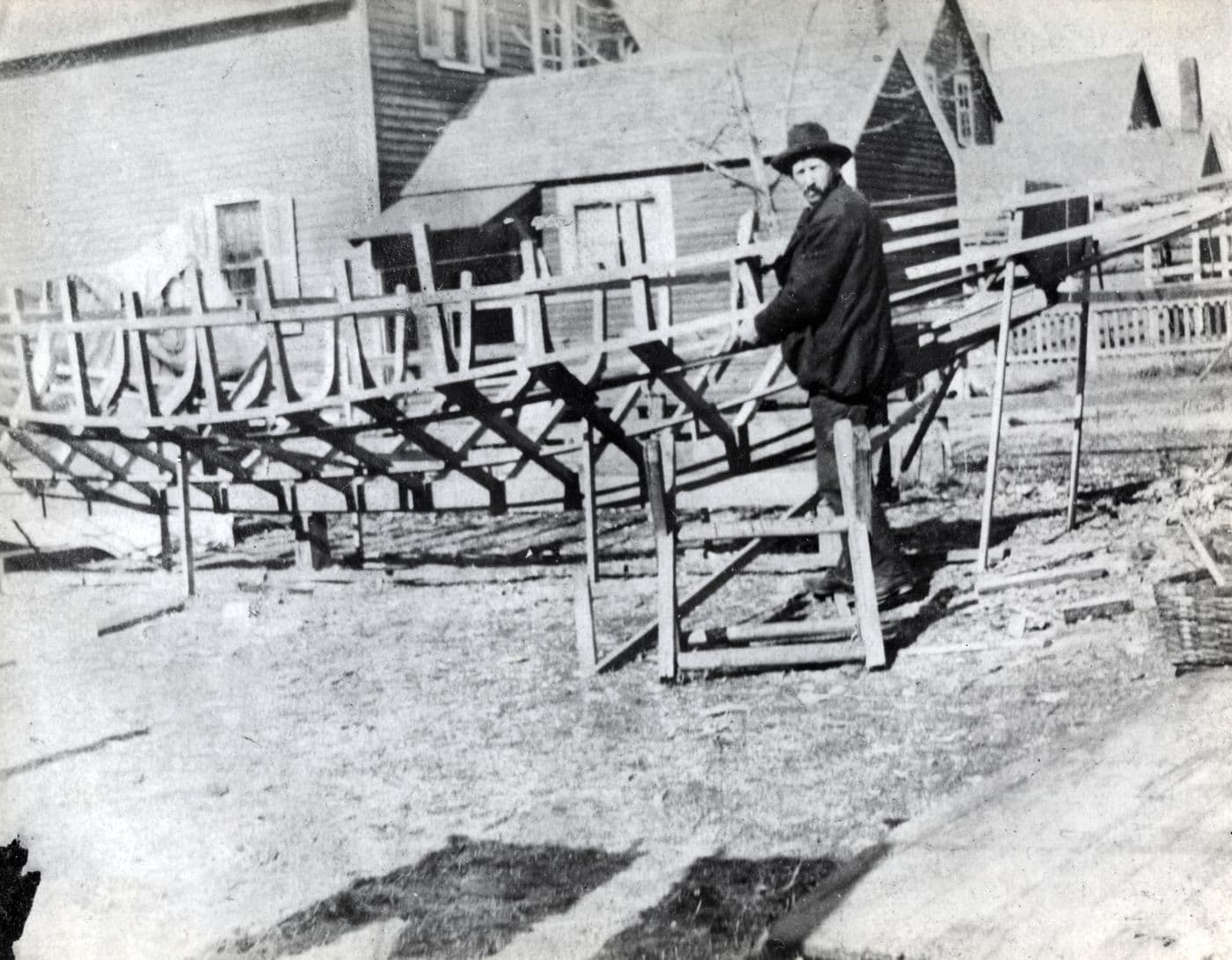Fred Miller standing beside the exposed wooden ribs of a catboat under construction in his backyard