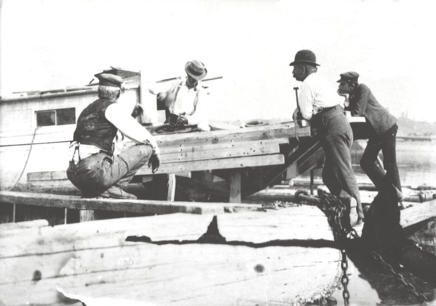Four men repairing a damaged wooden sloop near the Port Washington shoreline