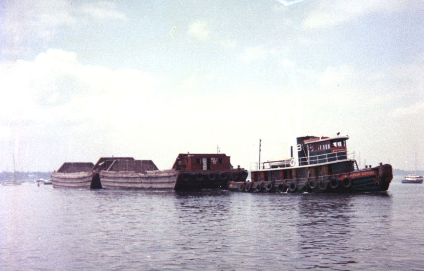 A tugboat moving a line of low sand barges through Manhasset Bay around 1965